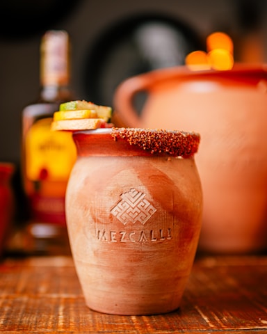 A rustic clay cup with the word 'Mezcally' embossed on its surface, garnished with slices of lime and other fruit on the rim, placed on a wooden table. In the blurred background, there is a bottle and another clay pot, creating a warm and inviting atmosphere.