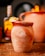 A rustic clay cup with the word 'Mezcally' embossed on its surface, garnished with slices of lime and other fruit on the rim, placed on a wooden table. In the blurred background, there is a bottle and another clay pot, creating a warm and inviting atmosphere.