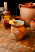 A rustic wooden table holds a traditional clay mug filled with a beverage, garnished with sliced citrus fruits on the rim. Beside it is a bottle of liquor, likely tequila. Other clay mugs and a large clay pot are visible in the background, suggesting a setting for enjoying drinks.