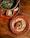 A vibrant and colorful spread featuring a rustic wooden table. A traditional Mexican dish is presented, with a taco filled with grilled meat, onions, jalapeños, and guacamole on a decorative terracotta plate with a dot pattern. Nearby, a large clay pot contains a grilled dish wrapped in banana leaves, accompanied by small bowls of guacamole, salsa, and a wedge of lime.