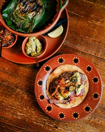 A vibrant and colorful spread featuring a rustic wooden table. A traditional Mexican dish is presented, with a taco filled with grilled meat, onions, jalape&ntilde;os, and guacamole on a decorative terracotta plate with a dot pattern. Nearby, a large clay pot contains a grilled dish wrapped in banana leaves, accompanied by small bowls of guacamole, salsa, and a wedge of lime.