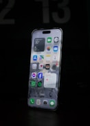 A modern smartphone is displayed with a lit-up screen showing various app icons, including weather, calendar, and social media applications, against a dark background. The screen's brightness highlights the device's sleek design.