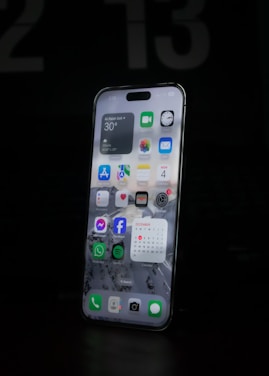 A modern smartphone is displayed with a lit-up screen showing various app icons, including weather, calendar, and social media applications, against a dark background. The screen's brightness highlights the device's sleek design.