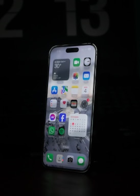 A modern smartphone is displayed with a lit-up screen showing various app icons, including weather, calendar, and social media applications, against a dark background. The screen's brightness highlights the device's sleek design.
