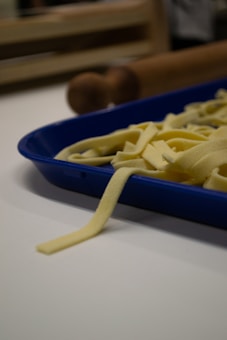 Freshly made pasta is laid out in a blue tray on a white countertop. In the background, there is a wooden rolling pin, slightly out of focus.