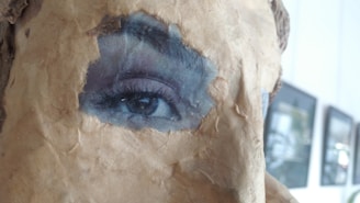 A close-up view of an artistic piece where a realistic eye is integrated into a textured surface resembling old, worn paper. The eye appears vivid and contrasts sharply with the surrounding material. In the background, blurred frames of art or photographs are visible on a wall.