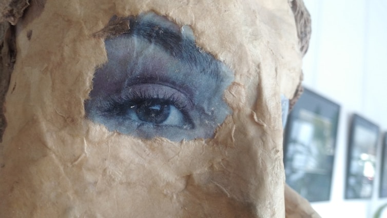 A close-up view of an artistic piece where a realistic eye is integrated into a textured surface resembling old, worn paper. The eye appears vivid and contrasts sharply with the surrounding material. In the background, blurred frames of art or photographs are visible on a wall.