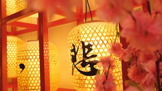 Large, ornate paper lanterns with intricate lattice patterns are illuminated, casting a warm golden glow. Black calligraphy is visible on the lanterns. Delicate pink cherry blossoms frame the scene, adding a touch of color and nature.