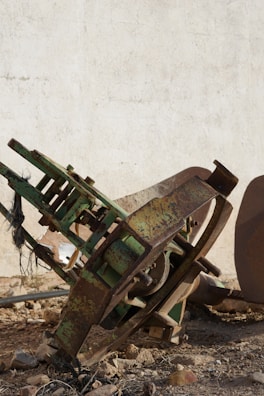 A rusted piece of heavy machinery lies on rocky ground against a worn, white wall. The machinery has visible signs of wear with peeling green paint and rusty metal components.