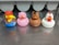 Four rubber duck toys are lined up on a metallic surface. Each duck is styled to resemble a different animal or character. The first duck wears a red cap and blue overalls, the second is painted pink to look like a pig, the third is brown with horse-like features, and the fourth is white with red details resembling a chicken.
