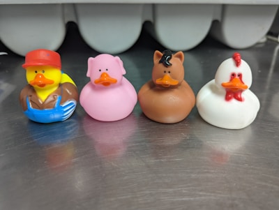 Four rubber duck toys are lined up on a metallic surface. Each duck is styled to resemble a different animal or character. The first duck wears a red cap and blue overalls, the second is painted pink to look like a pig, the third is brown with horse-like features, and the fourth is white with red details resembling a chicken.
