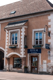 A rustic building with two large windows and a central door labeled 'Kolm Die Bäckerei', suggesting it is a bakery. The building has a stucco exterior with decorative stone elements and a traditional sloped roof. There are potted plants below the windows, which add a touch of greenery.