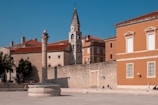 An ancient stone column stands in an open square next to a round stone structure. Surrounding the square are buildings with orange and beige facades, featuring red-tiled roofs. A bell tower with arched windows is visible in the background. Trees and a stone wall provide shade and texture to the scene, while a couple of people sit along the edge of the square.