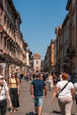 A bustling city street with people walking in both directions. The buildings on either side are tall and historic, featuring classic architectural details. The street leads to a medieval gate tower with a pointed roof and a small flag at the top, enhancing the historic ambience. The sky is clear and blue, suggesting a sunny day.
