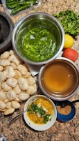 A variety of ingredients and dishes are spread out on a countertop, presumably for making an Indian snack. There are round, hollow puris piled together, fresh green chilies, bunches of cilantro, a lemon, and a tomato. Two metal bowls contain green spicy water and tamarind water. A small white bowl contains a mixture of chopped onions, chickpeas, and cilantro. A small blue bowl holds a powdered spice mix.
