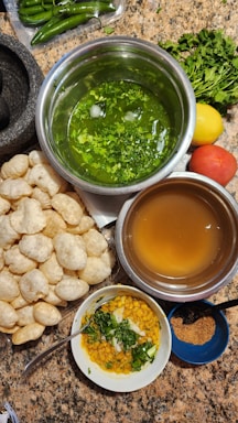 A variety of ingredients and dishes are spread out on a countertop, presumably for making an Indian snack. There are round, hollow puris piled together, fresh green chilies, bunches of cilantro, a lemon, and a tomato. Two metal bowls contain green spicy water and tamarind water. A small white bowl contains a mixture of chopped onions, chickpeas, and cilantro. A small blue bowl holds a powdered spice mix.