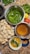A variety of ingredients and dishes are spread out on a countertop, presumably for making an Indian snack. There are round, hollow puris piled together, fresh green chilies, bunches of cilantro, a lemon, and a tomato. Two metal bowls contain green spicy water and tamarind water. A small white bowl contains a mixture of chopped onions, chickpeas, and cilantro. A small blue bowl holds a powdered spice mix.