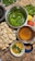A variety of ingredients and dishes are spread out on a countertop, presumably for making an Indian snack. There are round, hollow puris piled together, fresh green chilies, bunches of cilantro, a lemon, and a tomato. Two metal bowls contain green spicy water and tamarind water. A small white bowl contains a mixture of chopped onions, chickpeas, and cilantro. A small blue bowl holds a powdered spice mix.