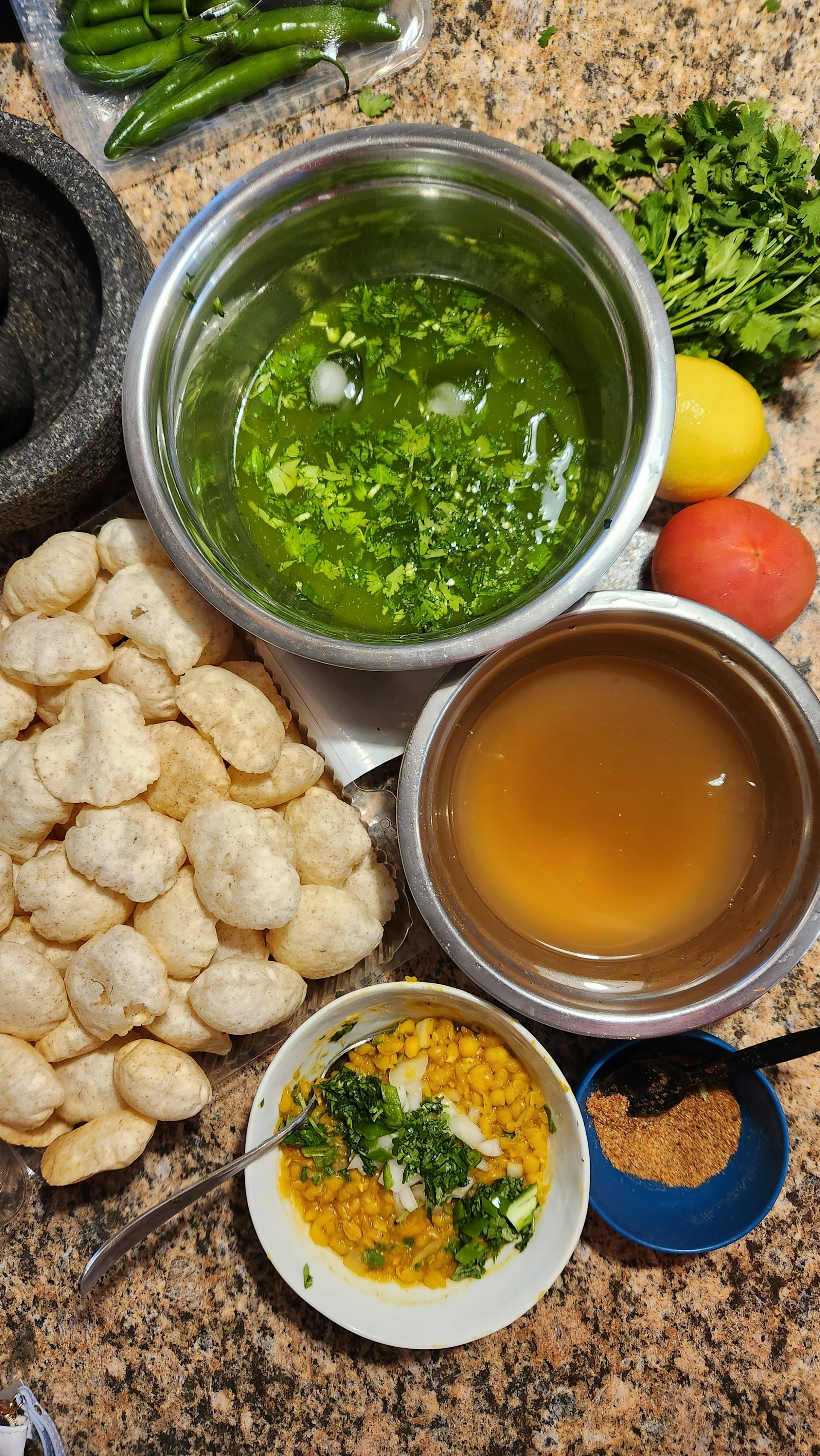A variety of ingredients and dishes are spread out on a countertop, presumably for making an Indian snack. There are round, hollow puris piled together, fresh green chilies, bunches of cilantro, a lemon, and a tomato. Two metal bowls contain green spicy water and tamarind water. A small white bowl contains a mixture of chopped onions, chickpeas, and cilantro. A small blue bowl holds a powdered spice mix.