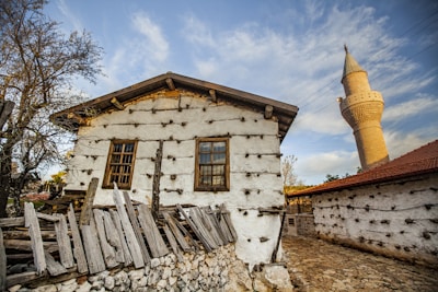 A traditional stone and wood house with a weathered wooden fence and a cobblestone pathway. The house has whitewashed walls with wooden beams and two windows at the front. A tall, beige minaret towers in the background. The setting includes leafless trees and some greenery, under a clear blue sky with wispy clouds.