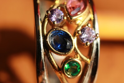A close-up view of a gold ring adorned with multiple gemstones. The gemstones include a large oval blue stone, two smaller round purple stones, and one round green stone. The setting features intricate gold work, adding elegance and detail to the piece.