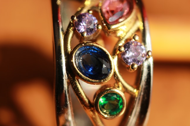 A close-up view of a gold ring adorned with multiple gemstones. The gemstones include a large oval blue stone, two smaller round purple stones, and one round green stone. The setting features intricate gold work, adding elegance and detail to the piece.