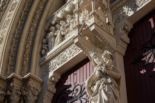 Intricate stone sculptures adorn a richly detailed gothic-style building facade. These sculptures include multiple figures, some of which appear to be religious or historical, intricately carved and positioned above arched doorways. The craftsmanship highlights delicate features and ornate architectural elements, casting shadows that add depth to the design.