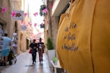 A narrow, sunlit street in a city adorned with pink flags and hanging signs. Several people walk along the street, two of them appearing to chat and hold items. On the right side, a bold yellow bag with text in French is prominently displayed in the foreground, while a street vendor and products are visible on the left.