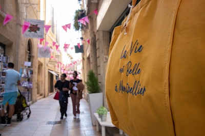 A narrow, sunlit street in a city adorned with pink flags and hanging signs. Several people walk along the street, two of them appearing to chat and hold items. On the right side, a bold yellow bag with text in French is prominently displayed in the foreground, while a street vendor and products are visible on the left.