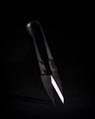 A pair of traditional black shears with sharp, overlapping blades positioned against a dark background. The lighting emphasizes the metallic sheen of the blades and the aged appearance of the handles.