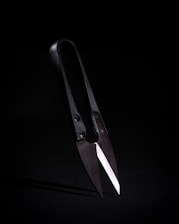 A pair of traditional black shears with sharp, overlapping blades positioned against a dark background. The lighting emphasizes the metallic sheen of the blades and the aged appearance of the handles.