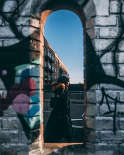 A person wearing a hooded coat with fur lining walks past a brick wall with colorful graffiti. The scene is framed by an arched opening in the wall, with a building and clear blue sky visible in the background.