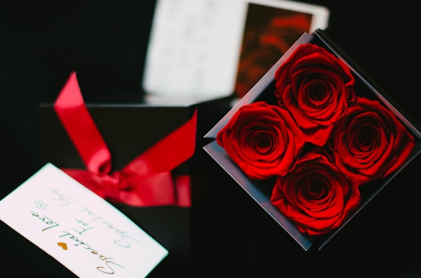 A box containing four vibrant red roses arranged neatly. Next to it, a black gift box with a red ribbon is partially visible. A card with the words 'Special Love' is also present, contributing to a romantic theme.