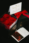 A close-up of a transparent box containing vibrant red roses sits prominently. In the background, there is a black gift box tied with a red ribbon. An elegant card with cursive writing is partially visible, adding a touch of personalization to the gift.