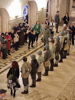 Several people, some in historical military uniforms with helmets, are standing in an indoor setting resembling a museum or historical building. They are being observed by a group of people, some with cameras and phones capturing the moment. The scene takes place in a grand hall with marble floors and arches, and there is a feeling of anticipation or ceremony in the air.