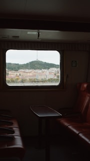 A view through a train or ferry window revealing a cityscape with a prominent hill and a transmission tower in the distance. Inside, there are red leather seats and a small table creating a cozy setting.