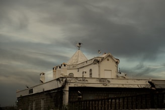 A building with a white, triangular roof adorned with several seagulls perched on various edges. The structure, appearing slightly weathered, shows antennas and a satellite dish. The sky is filled with dark, moody clouds, casting a gloomy atmosphere over the scene.