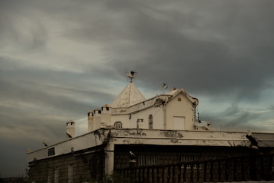 A building with a white, triangular roof adorned with several seagulls perched on various edges. The structure, appearing slightly weathered, shows antennas and a satellite dish. The sky is filled with dark, moody clouds, casting a gloomy atmosphere over the scene.