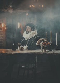 A man in period costume with a ruffled collar sits at a wooden table in a dimly lit, atmospheric setting. The scene includes a candelabra with lit candles, a bottle, a glass chalice, and a bowl of fruit, suggesting a historical or theatrical theme. The background is hazy, creating a moody and dramatic ambience.