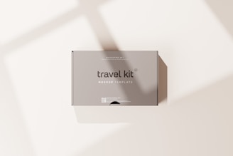 A minimalist travel kit box with the words 'travel kit' and 'mockup template' printed on it. The box is placed on a light surface with soft shadows cast across it.