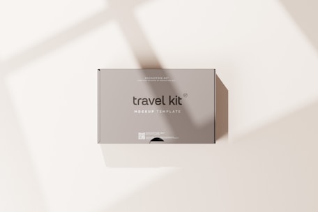 A minimalist travel kit box with the words 'travel kit' and 'mockup template' printed on it. The box is placed on a light surface with soft shadows cast across it.