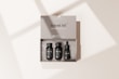 A beige travel kit box containing three small, black bottles neatly arranged inside. The kit includes items labeled as shampoo, conditioner, and a vitamin C serum. The minimalist packaging design is simple and elegant, with the words 'travel kit' and 'mockup template' clearly visible on the box. Soft shadows are cast on the surface, creating a subtle, natural lighting effect.