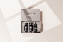 A beige travel kit box containing three small, black bottles neatly arranged inside. The kit includes items labeled as shampoo, conditioner, and a vitamin C serum. The minimalist packaging design is simple and elegant, with the words 'travel kit' and 'mockup template' clearly visible on the box. Soft shadows are cast on the surface, creating a subtle, natural lighting effect.