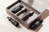 The image features a minimalist packaging design for a travel kit. There is an open cardboard box displaying slots for containers, accompanied by three black bottles with labels. Two bottles have dropper caps while the third has a regular cap.
