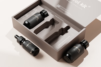 The image features a minimalist packaging design for a travel kit. There is an open cardboard box displaying slots for containers, accompanied by three black bottles with labels. Two bottles have dropper caps while the third has a regular cap.