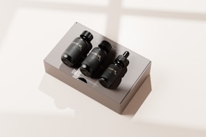 A set of three black bottles with droppers labeled as a travel kit, placed neatly on a rectangular, reflective, light-colored box. The scene is bathed in soft natural light, casting gentle shadows.