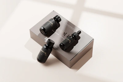 A set of black cosmetic dropper bottles displayed on a light-colored surface. Three bottles are placed on top of a rectangular box, while one bottle stands beside it. The box and bottles have minimalist labels with text and design elements. Light from a window creates soft shadows across the scene.