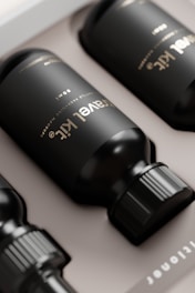 Several black bottles laying horizontally in a box. The bottles have a sleek, modern design with a matte finish and labeled with gold text. The box they are placed in is a light beige color, providing contrast to the dark bottles.