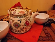 An ornately decorated ceramic teapot with floral and bird designs rests on a red napkin placed on a wooden table. It has a bamboo handle and is surrounded by white bowls, one of which contains a spoon. A plate of cookies sits nearby, adding a cozy touch.