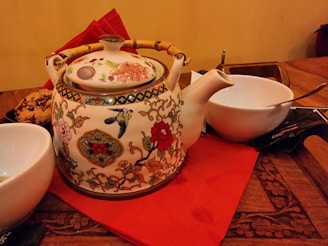 An ornately decorated ceramic teapot with floral and bird designs rests on a red napkin placed on a wooden table. It has a bamboo handle and is surrounded by white bowls, one of which contains a spoon. A plate of cookies sits nearby, adding a cozy touch.